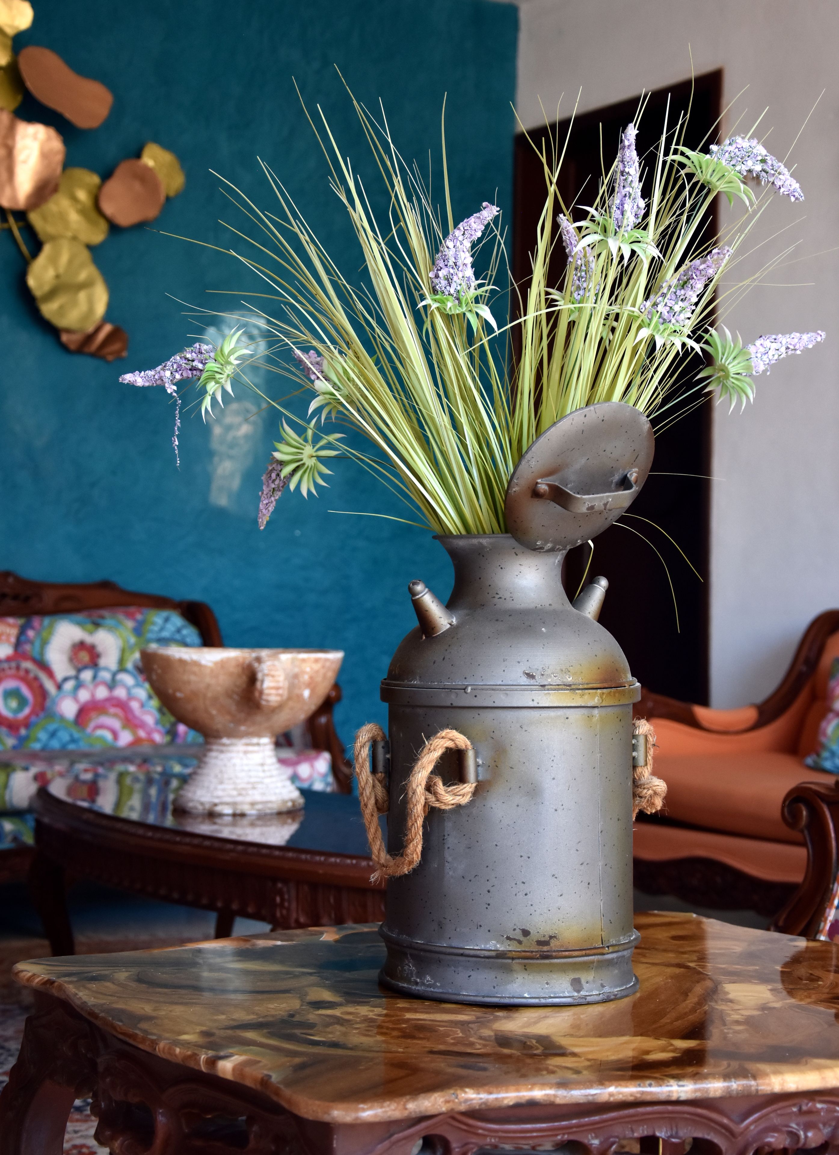 A rustic metal vase with a rope handle on a wooden table, filled with green plants and purple flowers.