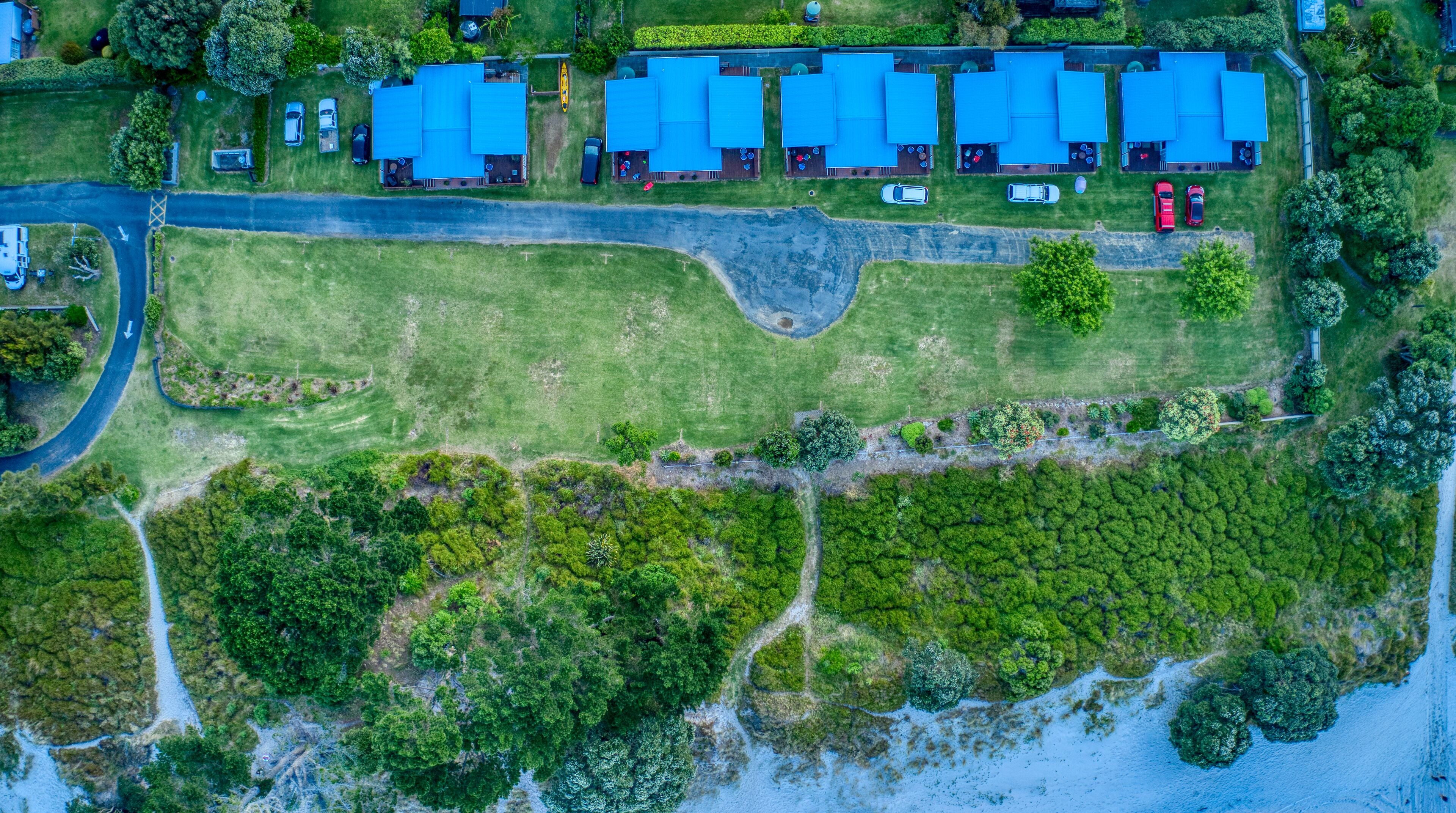 An aerial view of a residential area with multiple blue-roofed buildings, a winding road, and a dense green forest area.