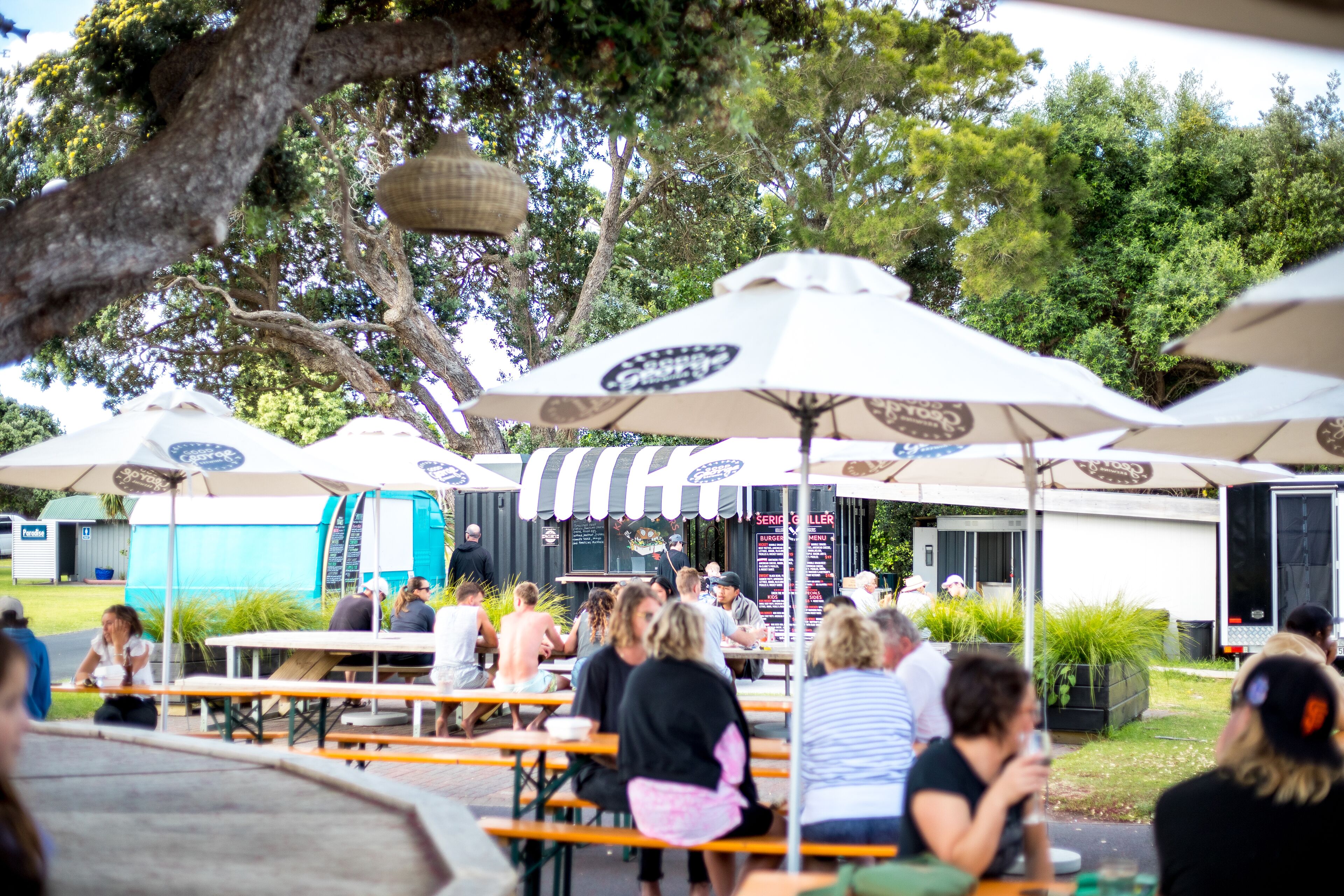 An outdoor dining area with tables and umbrellas, surrounded by trees and a building with a sign.