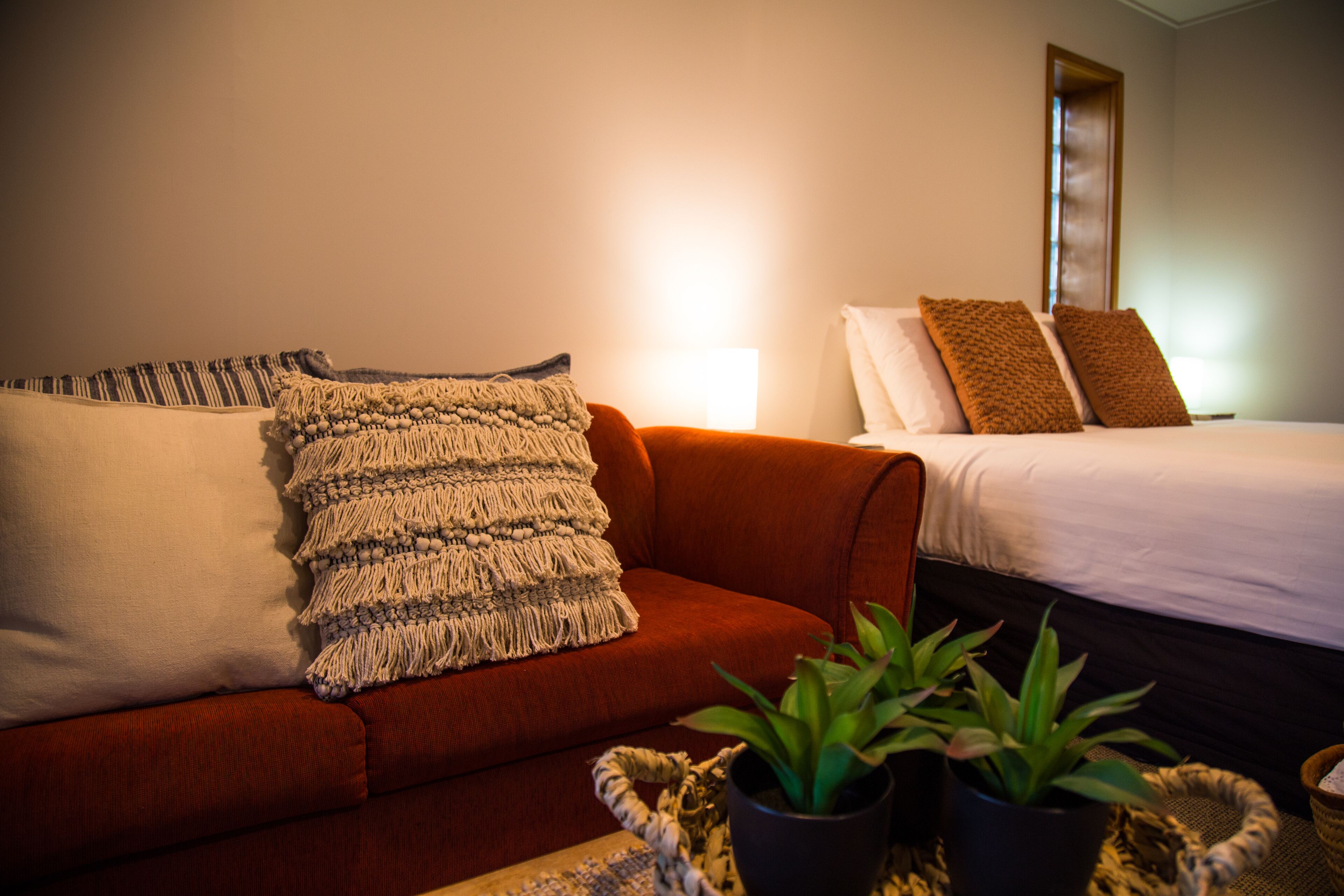 A hotel room with a red sofa, a bed with white bedding, and two potted plants on a side table.