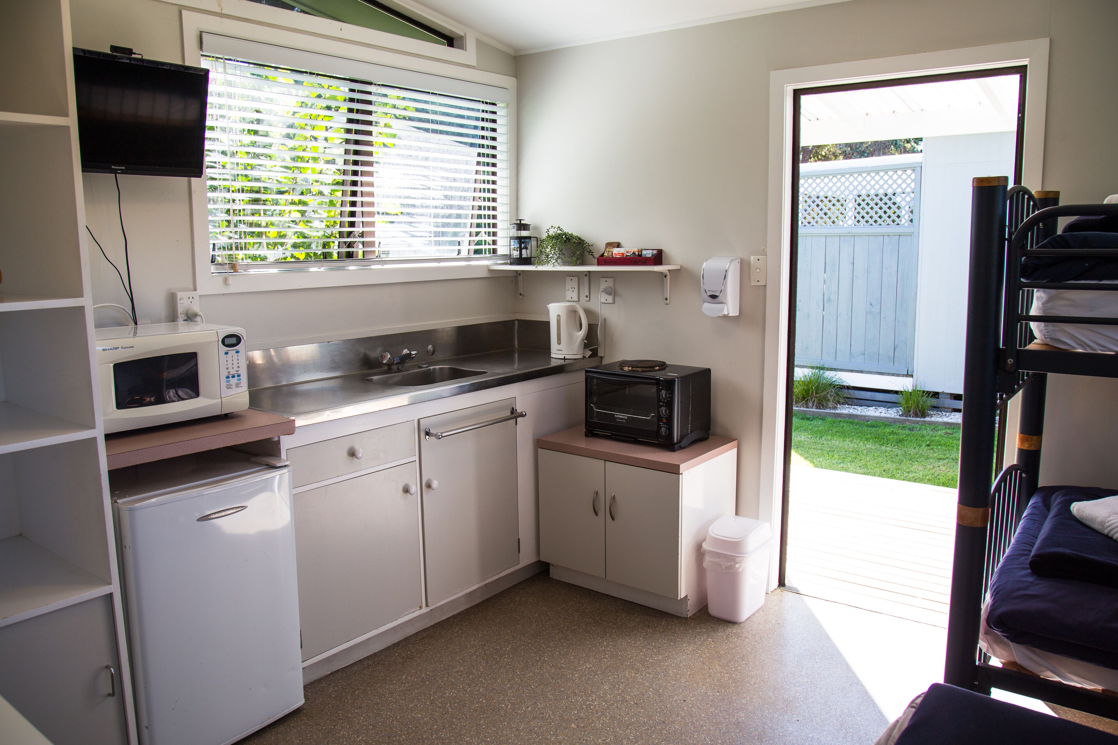 A compact kitchen with a microwave, kettle, and sink. There is a small refrigerator and a window with blinds. A bunk bed is visible in the background.