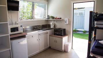 Een compacte keuken met een magnetron, waterkoker en spoelbak. Er is een kleine koelkast en een raam met zonwering. Achterin is een stapelbed zichtbaar.