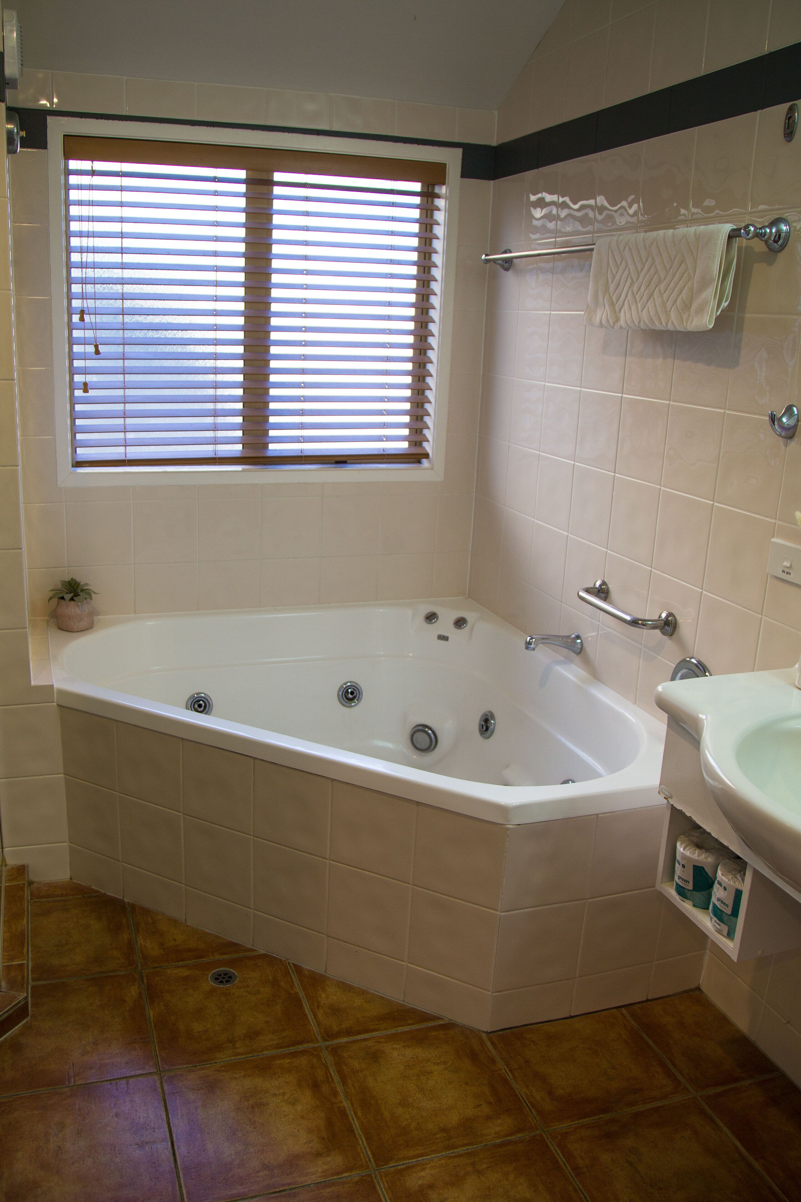 Een badkamer met een bad, een raam met zonwering, een handdoekrek en een wastafel.