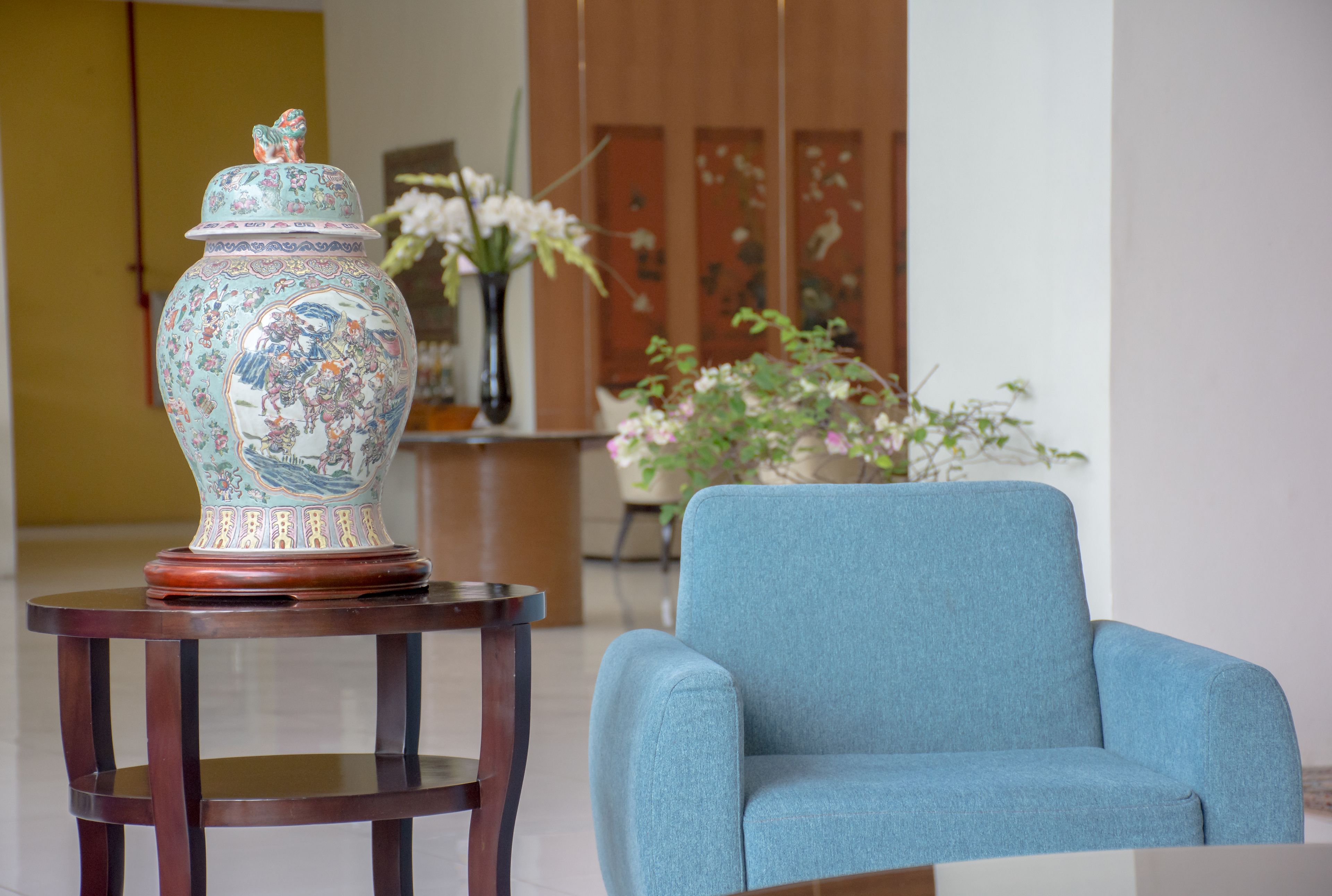 A blue armchair and a decorative vase on a wooden table in a room with a wooden cabinet and a vase of flowers.