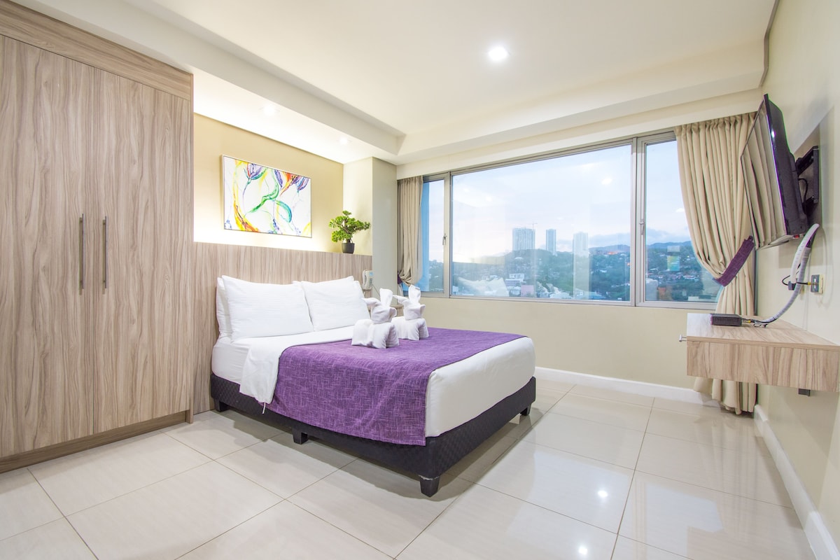 A hotel room with a large bed, a city view, a TV, and a wooden wardrobe.