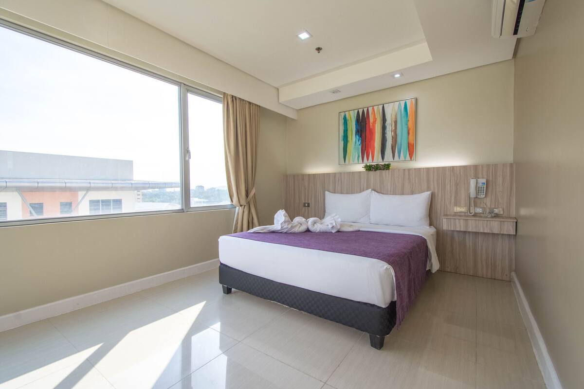 A hotel room with a large bed, a window with curtains, a wall-mounted phone, and a city view.