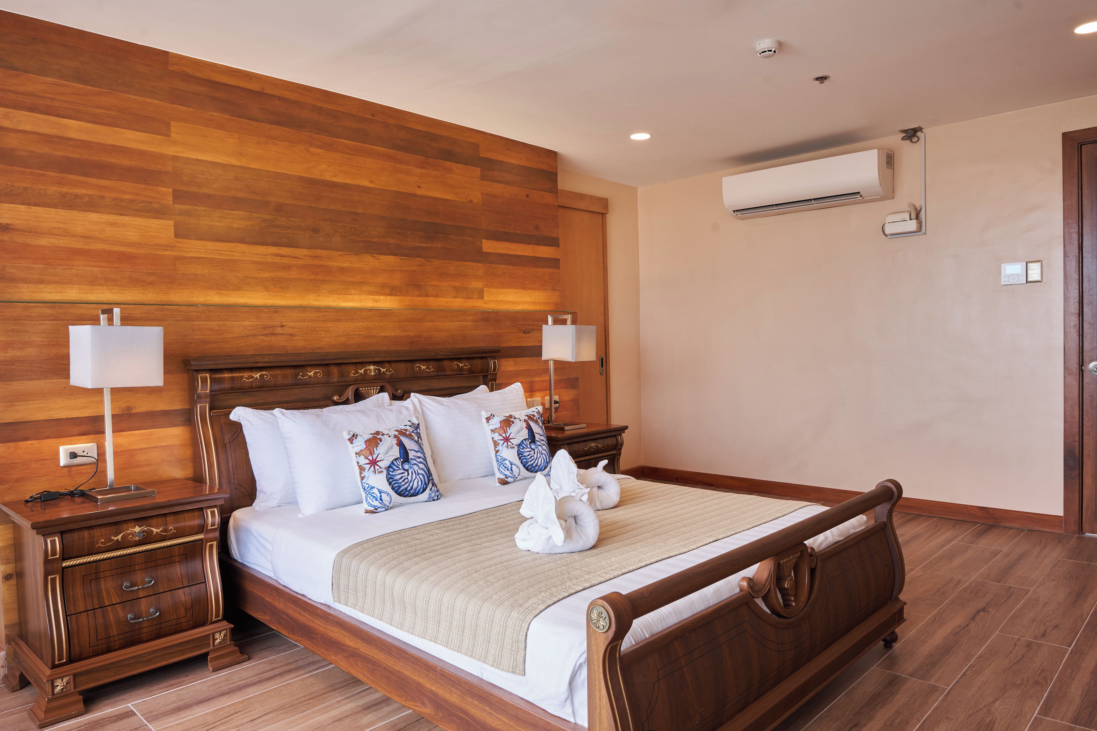 A hotel room with a wooden headboard, a bed with white linens, decorative pillows, a bedside table with a lamp, and an air conditioning unit on the wall.