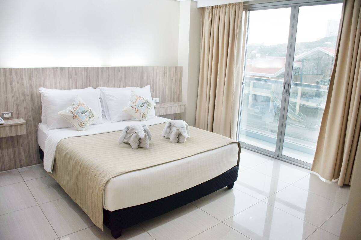 A hotel room with a large bed, two bedside tables, a window with curtains, and a view of buildings outside.