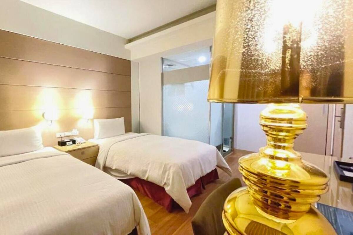 A hotel room with two beds, a nightstand, a lamp, and a view of the outside.