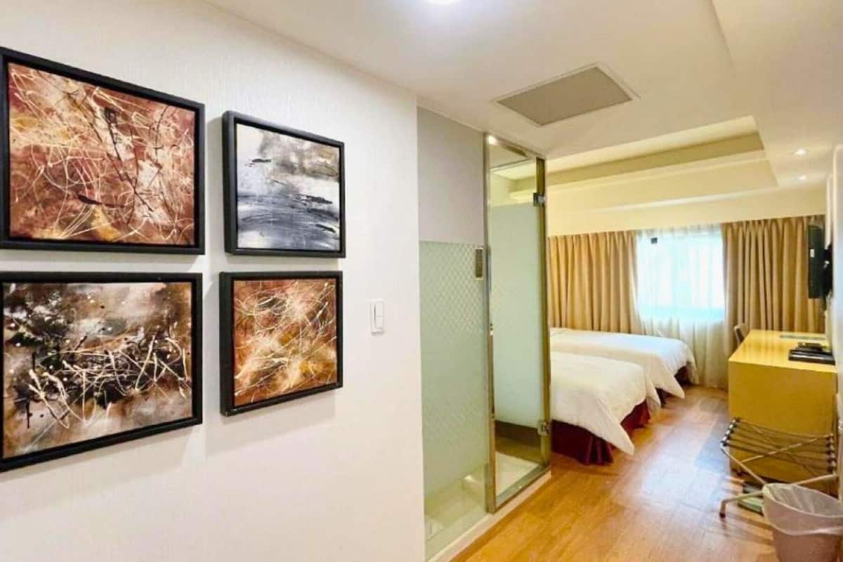 A hotel room with four abstract paintings on the wall, a bed, a desk with a chair, and a small trash bin.