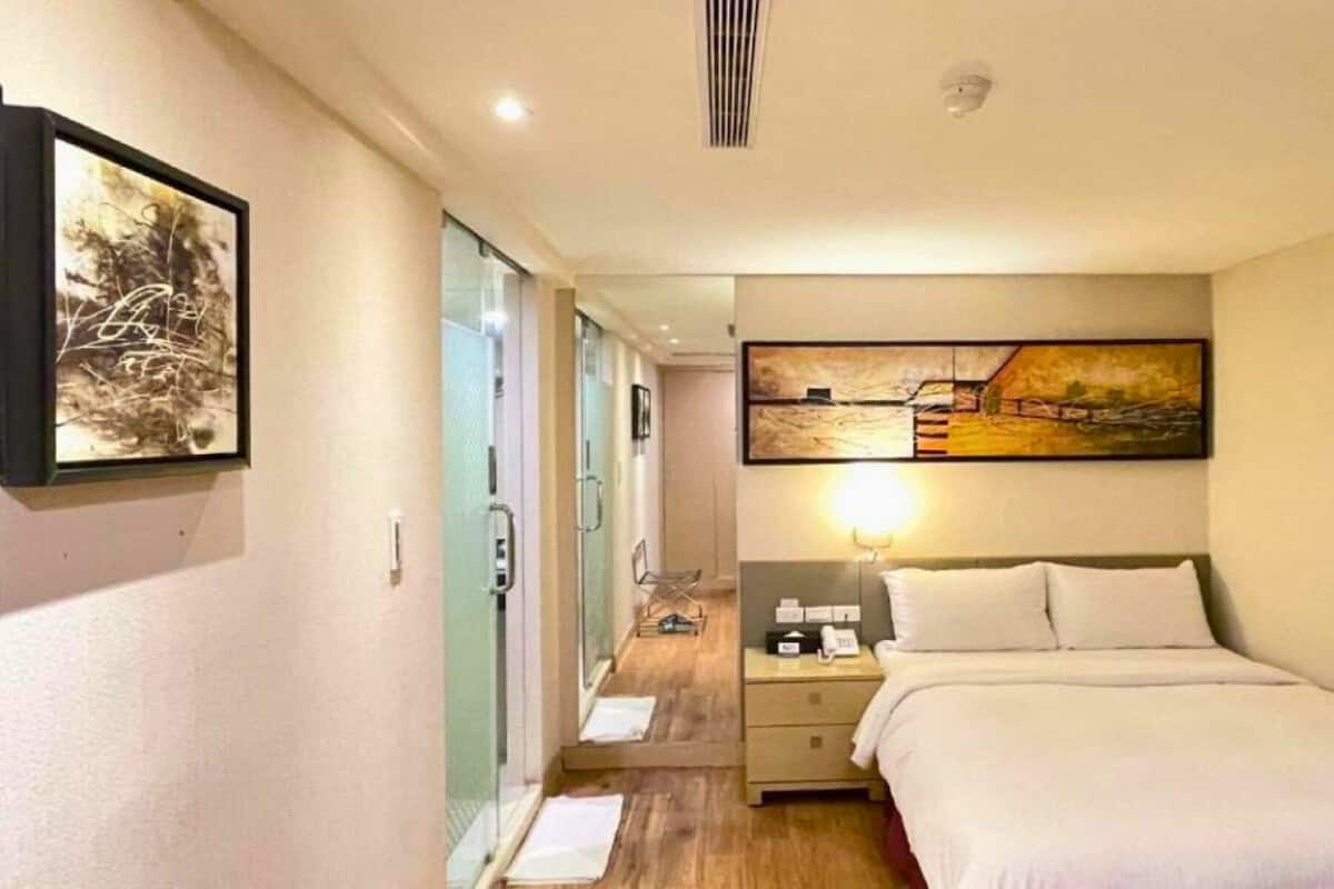 A hotel room with a bed, a bedside table, a nightstand, a lamp, a painting on the wall, and a door leading to a bathroom.