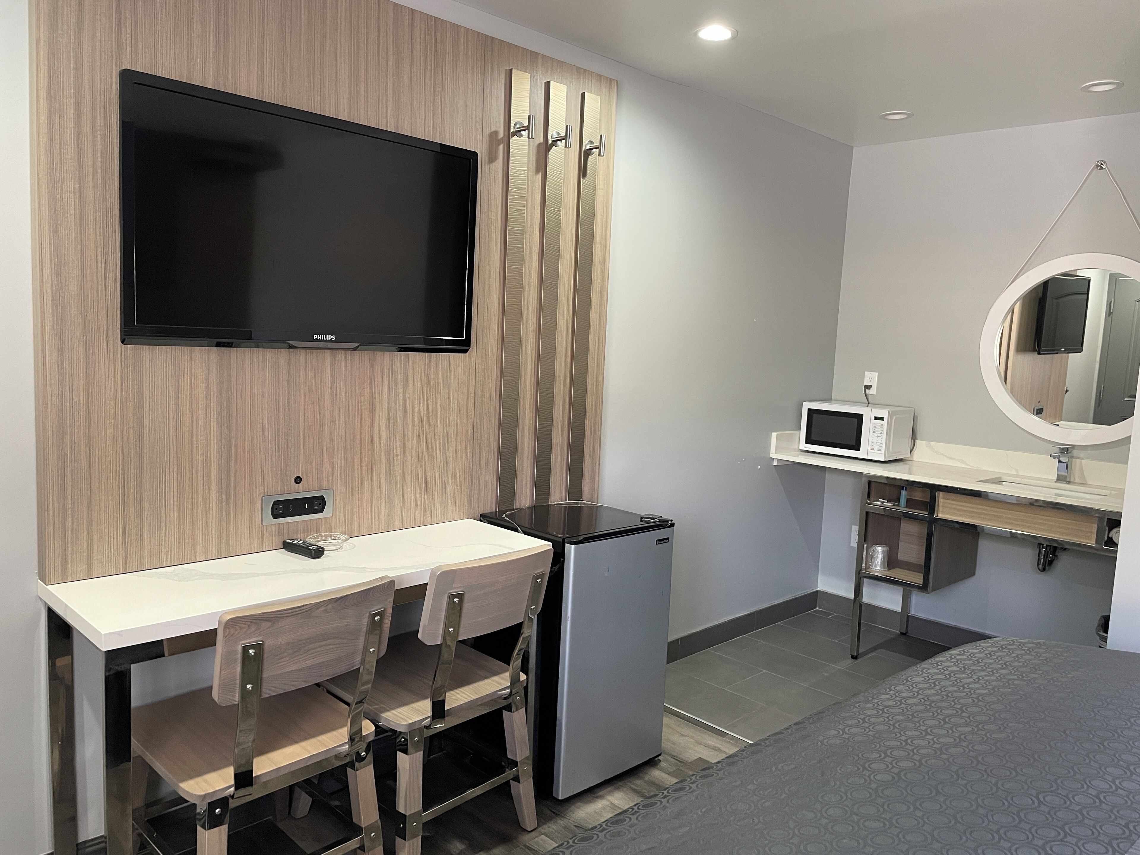 A compact hotel room with a wooden wall, a flat-screen TV, a refrigerator, a microwave, a sink, and a mirror.