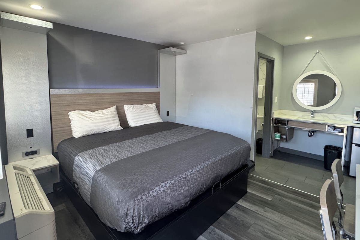 Quarto moderno com uma cama grande, escrivaninha embutida e banheiro visível através de uma porta aberta.