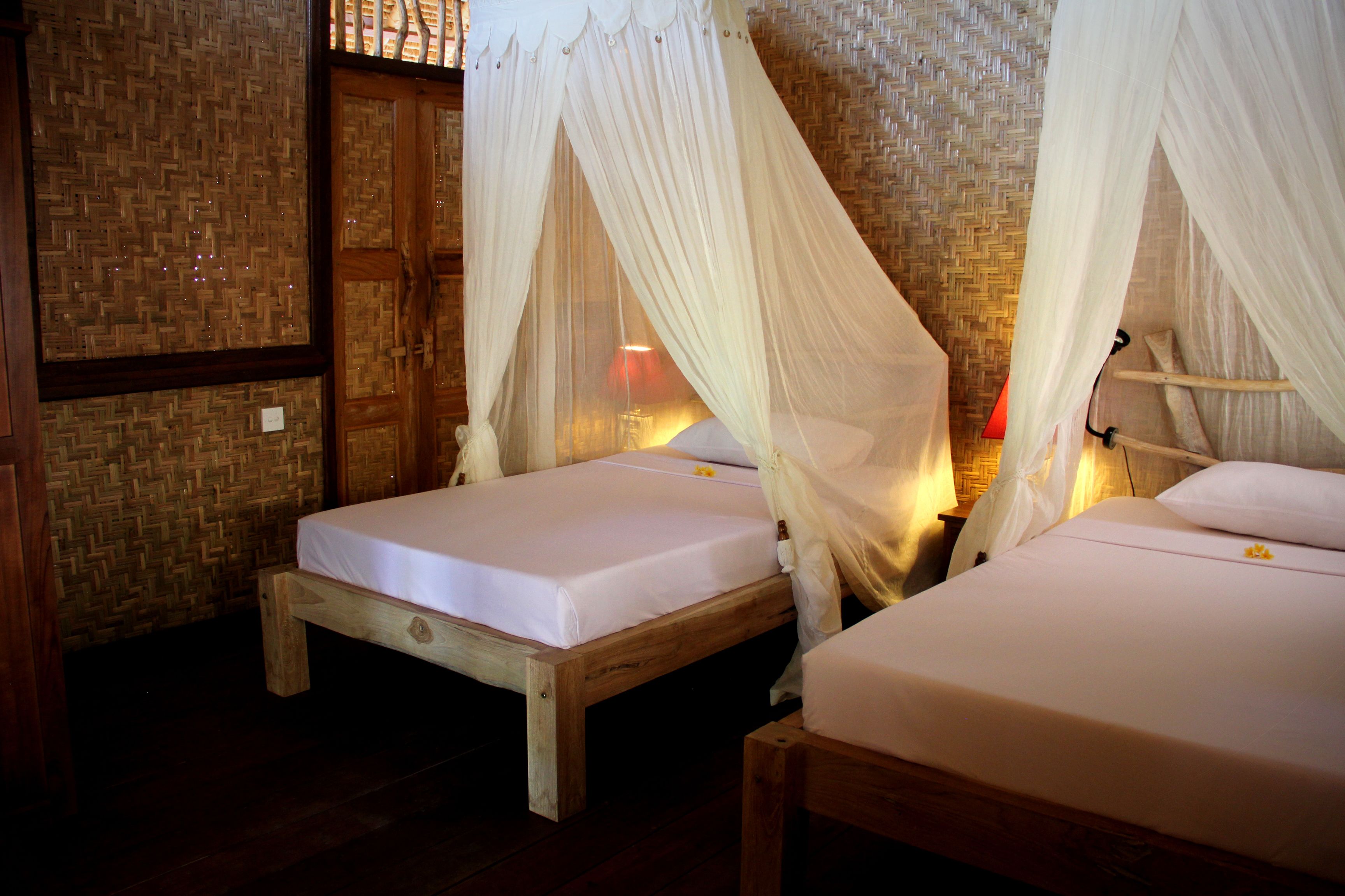 Two wooden beds with white bedding and mosquito netting, a wooden door, and a woven wall.