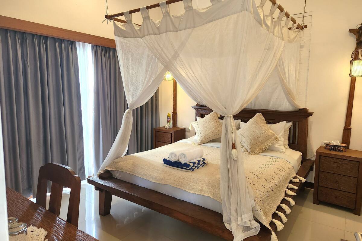 A four-poster canopy bed with a wooden headboard, a bedside table with a lamp, and a chair at the foot of the bed.
