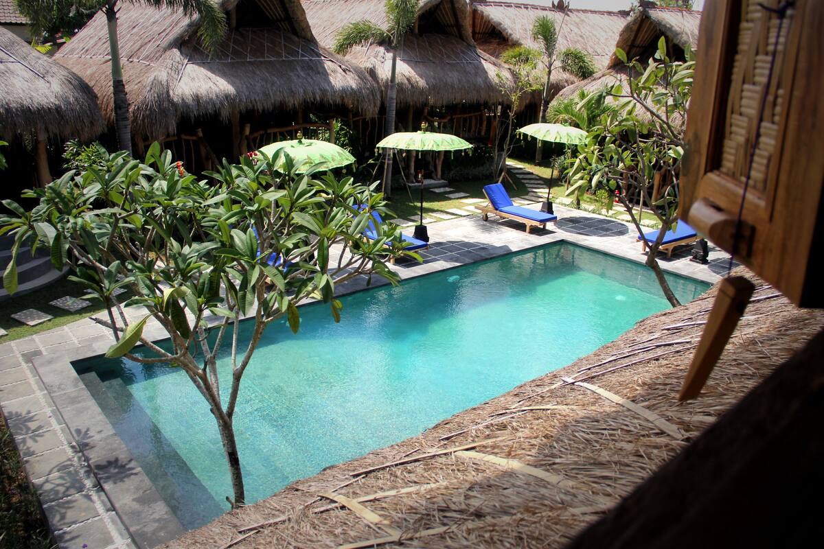A swimming pool with lounge chairs and umbrellas, surrounded by thatched-roof huts.