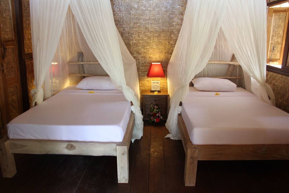 Two wooden beds with white linens and canopy beds, a nightstand with a red lamp, and a woven wall.