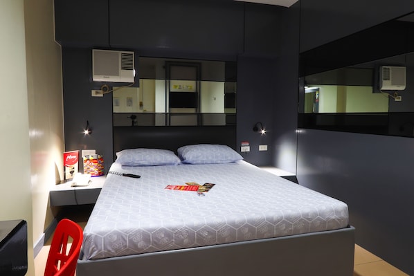 Una habitación de hotel con una cama, un escritorio con silla, un espejo y un equipo de aire acondicionado.