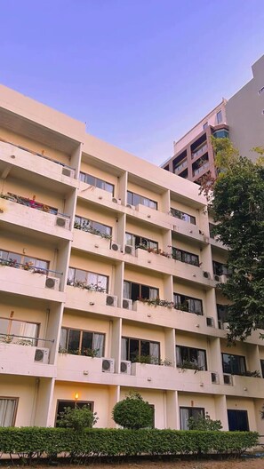 Un immeuble résidentiel à plusieurs étages, doté de balcons, de climatiseurs et d’une haie bien entretenue à l’avant.