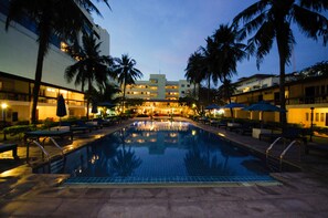 Une piscine entourée de palmiers et de bâtiments d’hôtel, avec des chaises longues et des parasols.