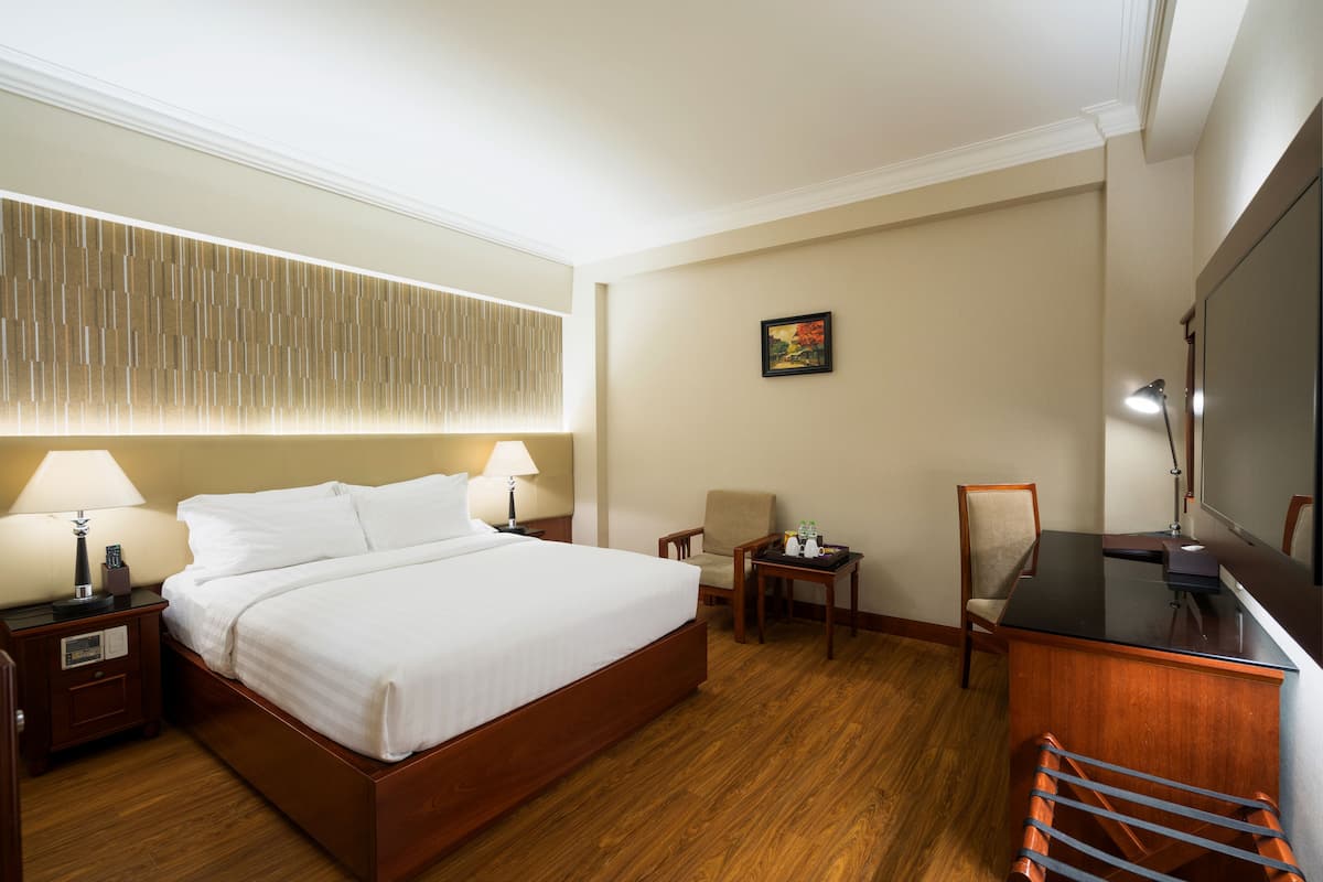 A hotel room with a large bed, two bedside tables with lamps, a desk with a chair, a TV, and a wooden floor.