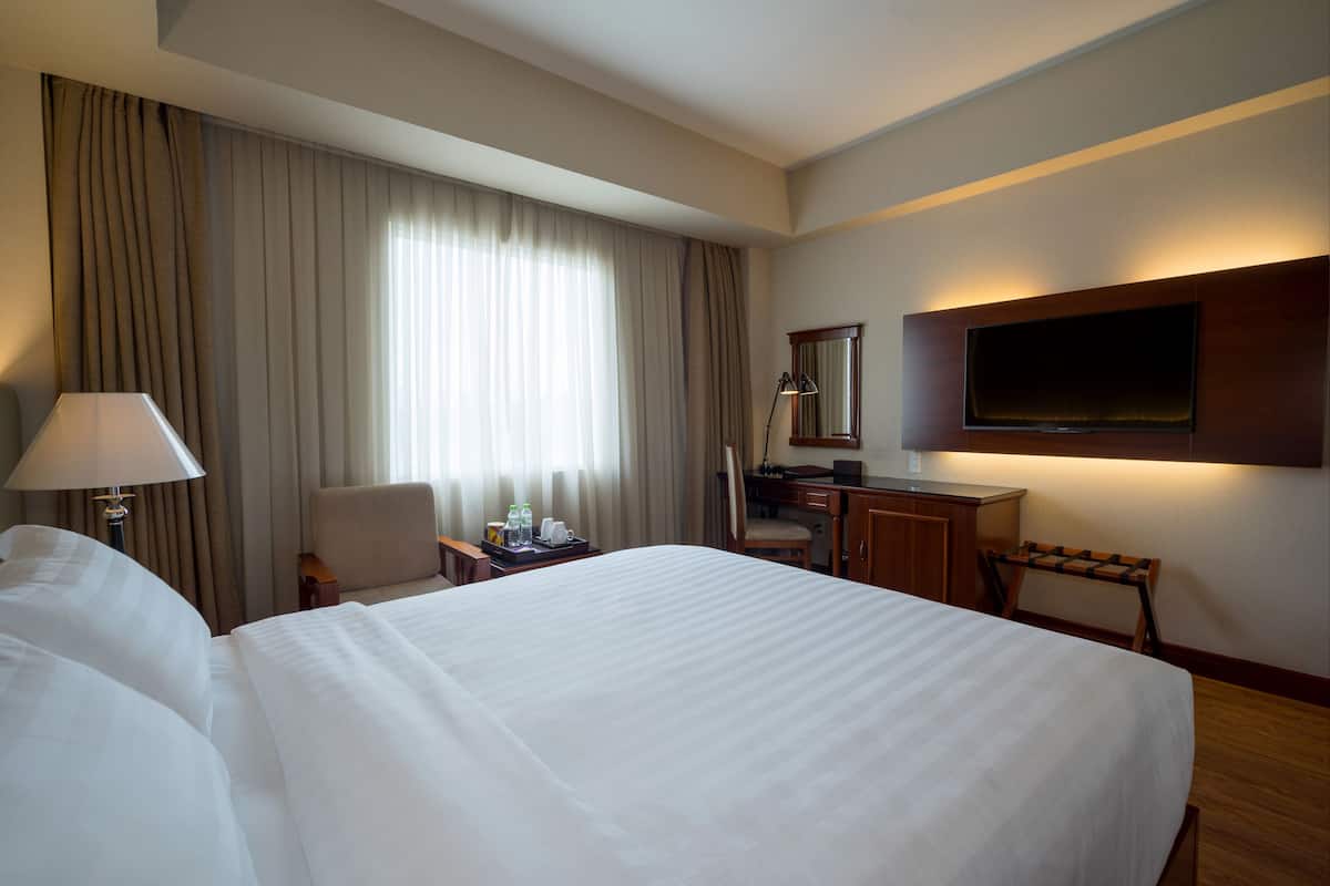 A hotel room with a large bed, a desk, a chair, a TV mounted on the wall, and a window with curtains.