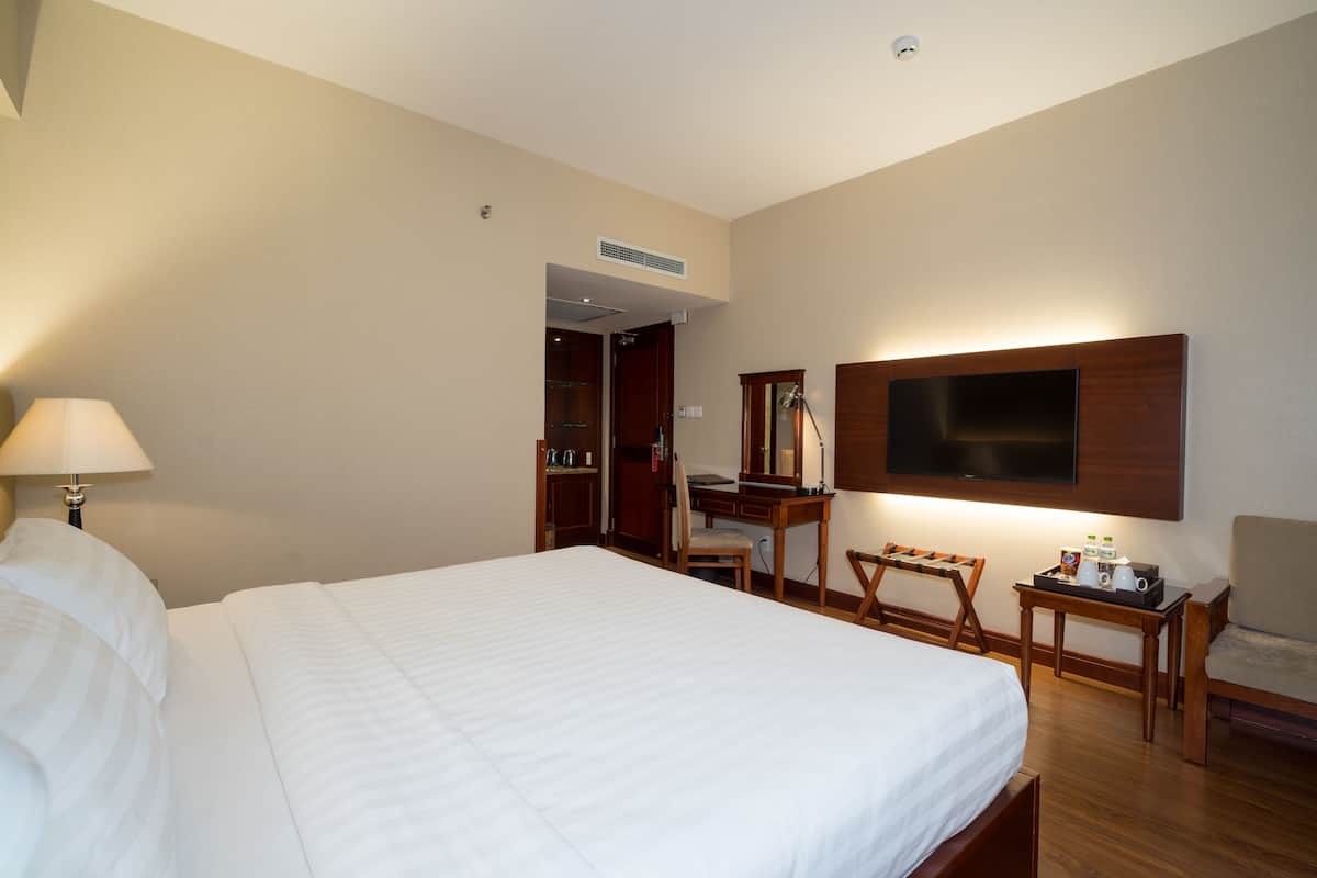 A hotel room with a large bed, a desk, a chair, a TV mounted on the wall, and a small table with a lamp.