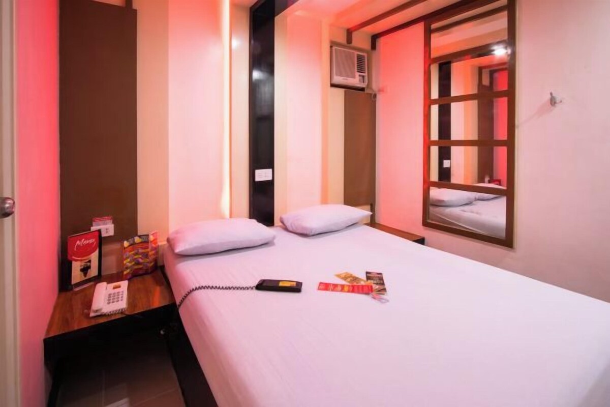 A hotel room with a bed, a bedside table with a phone and a book, a window with a view, and a wall-mounted air conditioner.