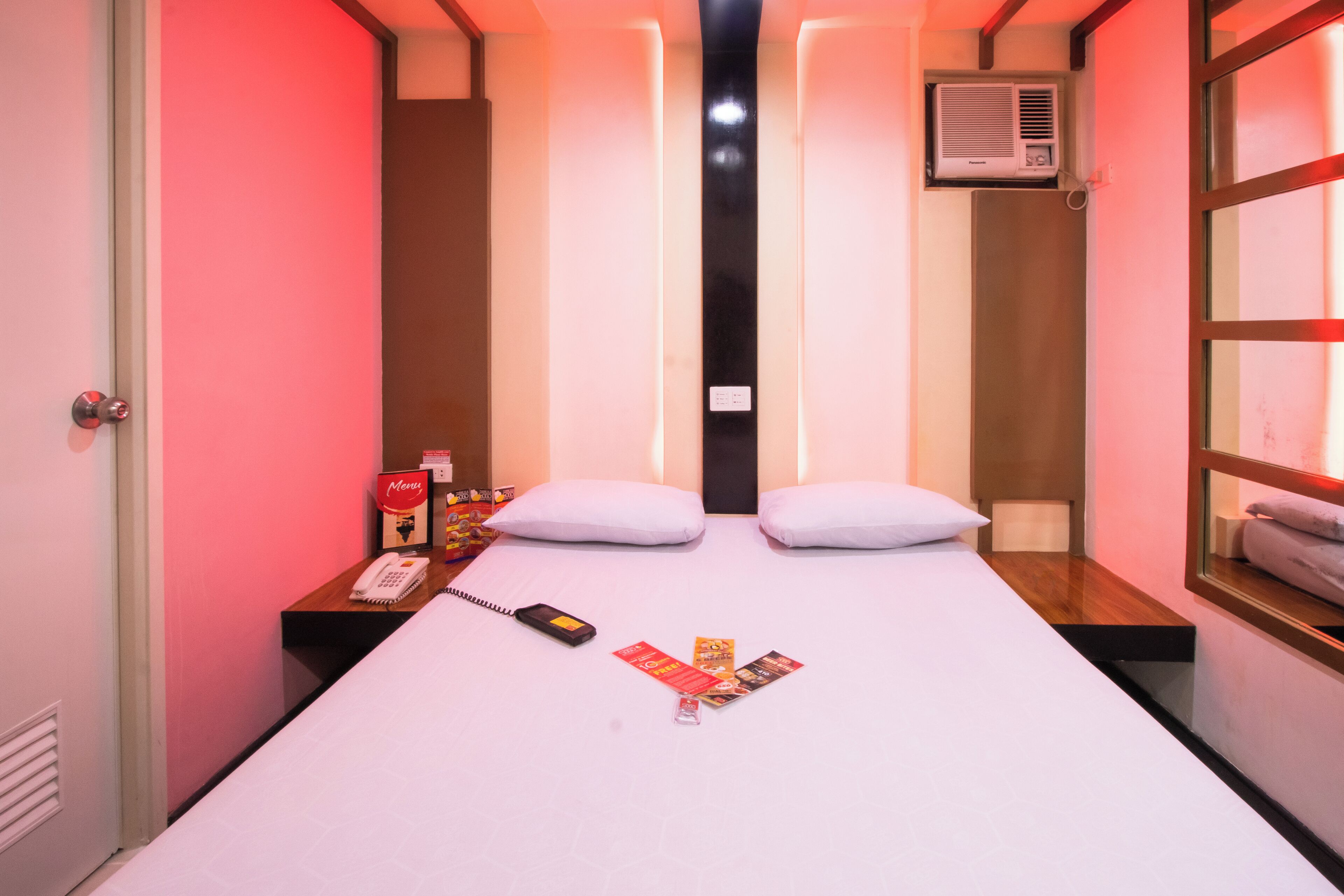 A hotel room with a bed, two pillows, a bedside table with a phone and magazines, a window with a towel, and an air conditioning unit.