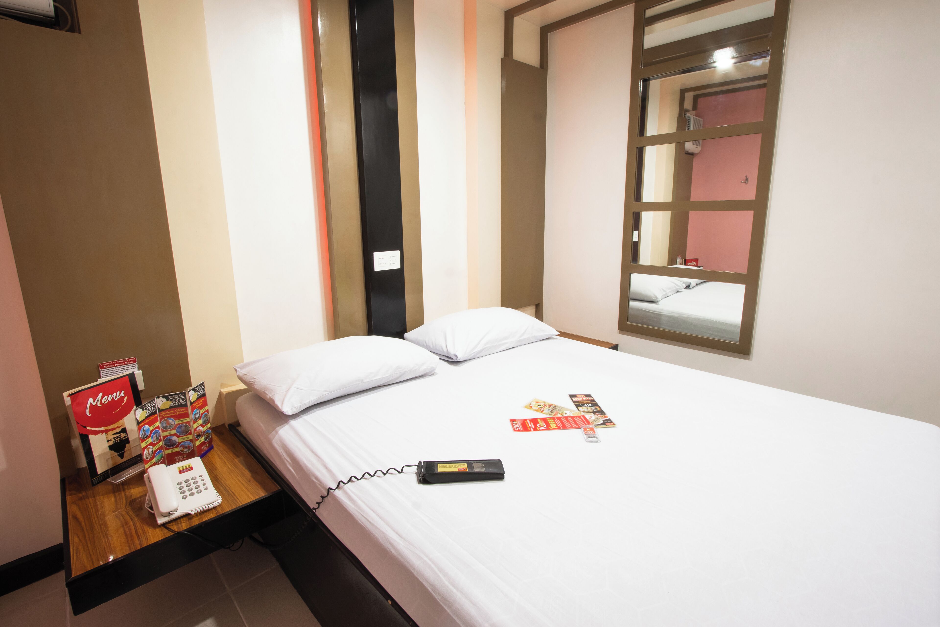 Habitación de hotel con cama, mesita de noche con teléfono, atril para el menú y un espejo.