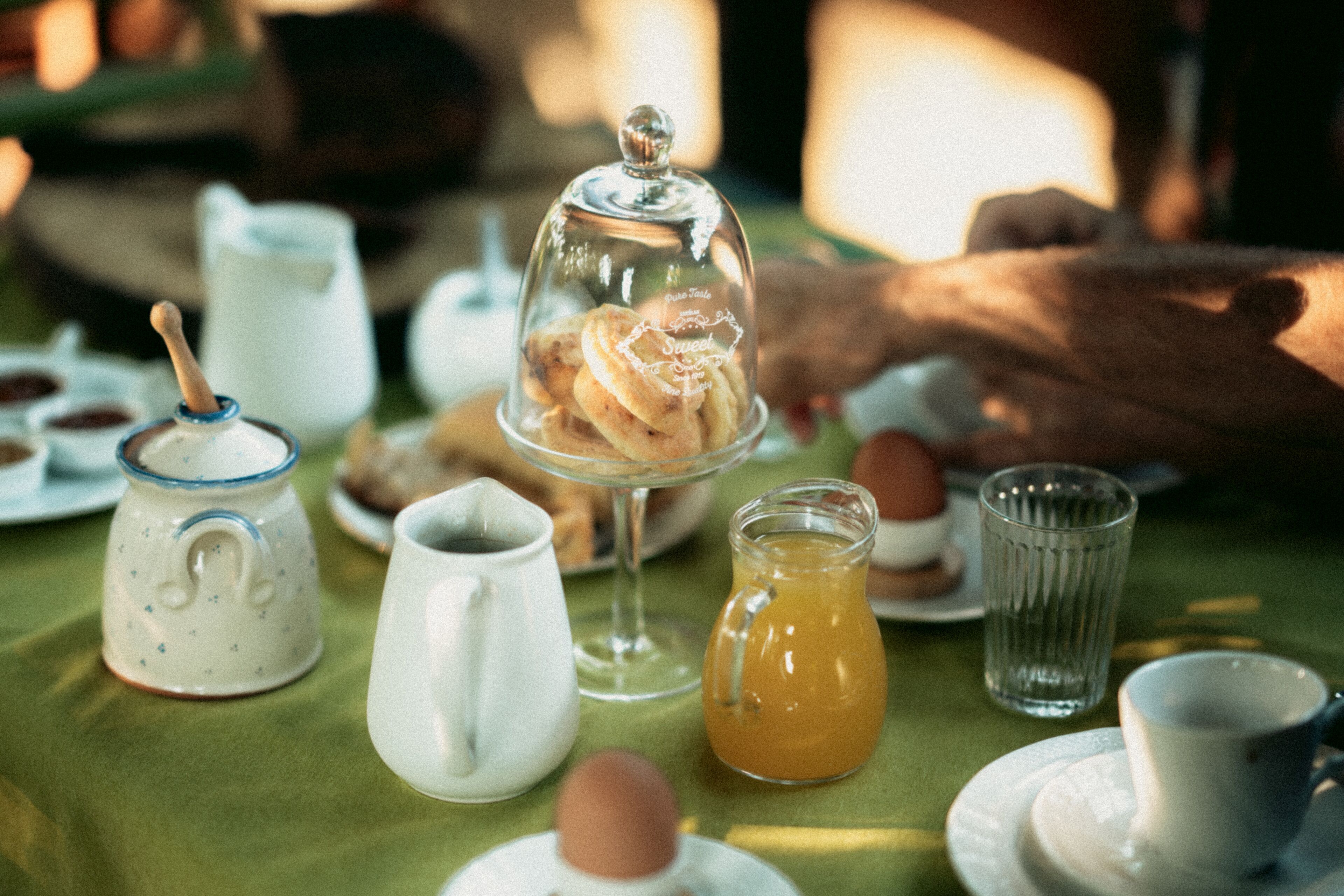 Een ontbijttafel met diverse spullen, zoals een karaf, een beker, een glas en een afgedekte schaal met gebak.