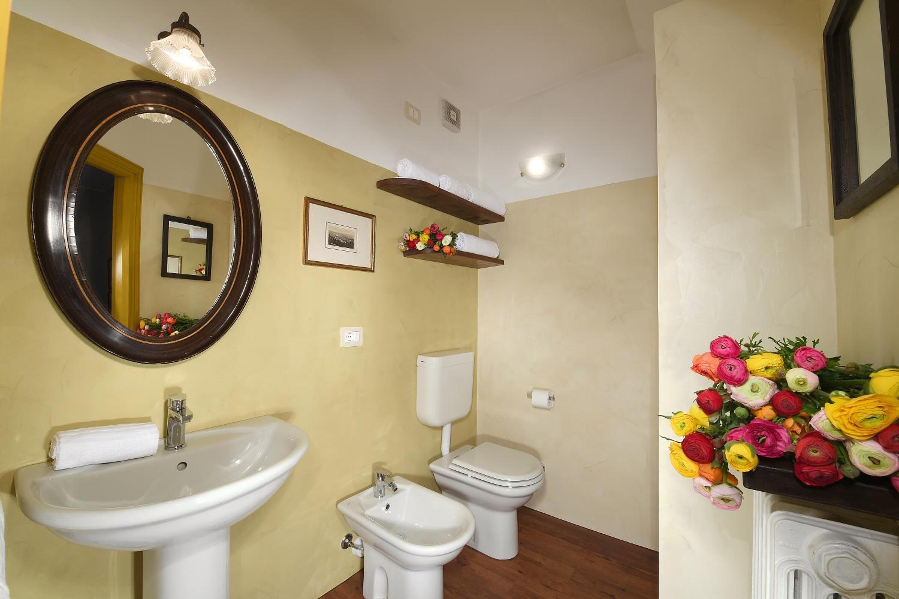 Een badkamer met een wastafel, toilet, bidet en een spiegel. Er staan bloemstukken op de wastafel en een plank boven het toilet.
