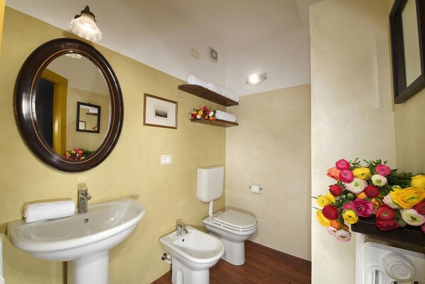A bathroom with a sink, bidet, toilet, and a mirror. There are flower arrangements on the counter and a shelf above the toilet.