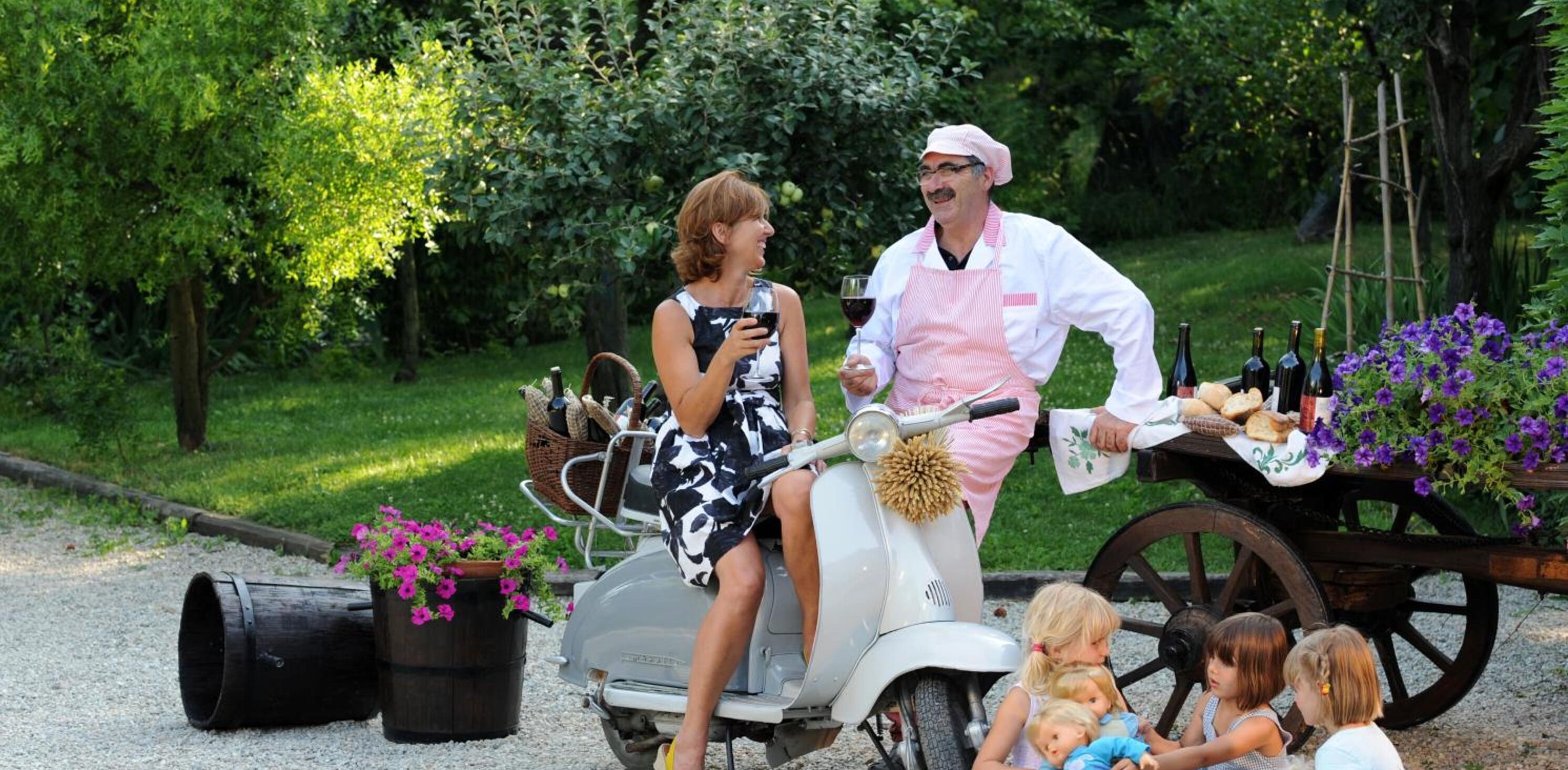 Una donna su uno scooter con un uomo in abiti da chef, un carretto con cibo e bevande, e dei bambini seduti a terra.