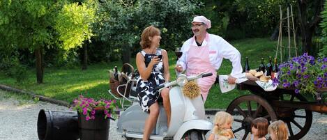 A woman on a scooter with a man in chef attire, a cart with food and drinks, and children sitting on the ground.
