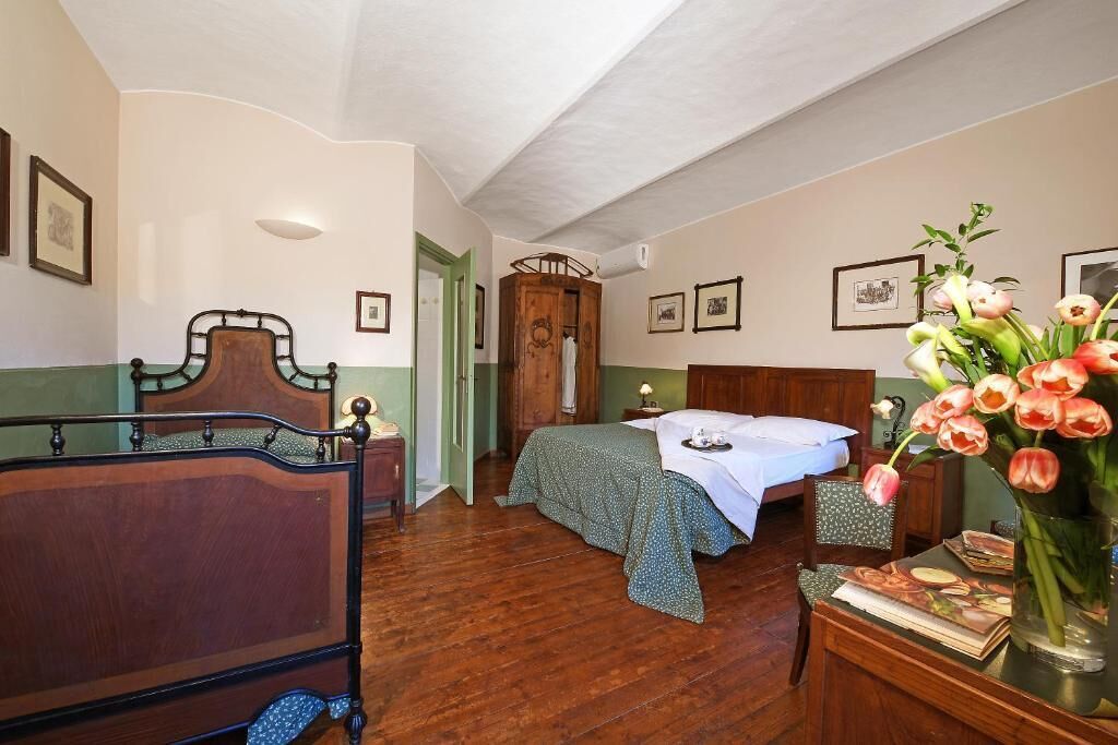 Une pièce comprenant un lit en bois, un plus petit lit recouvert d’une couverture verte, une armoire en bois, une table de chevet avec une lampe et un vase rempli de fleurs posé sur une table.