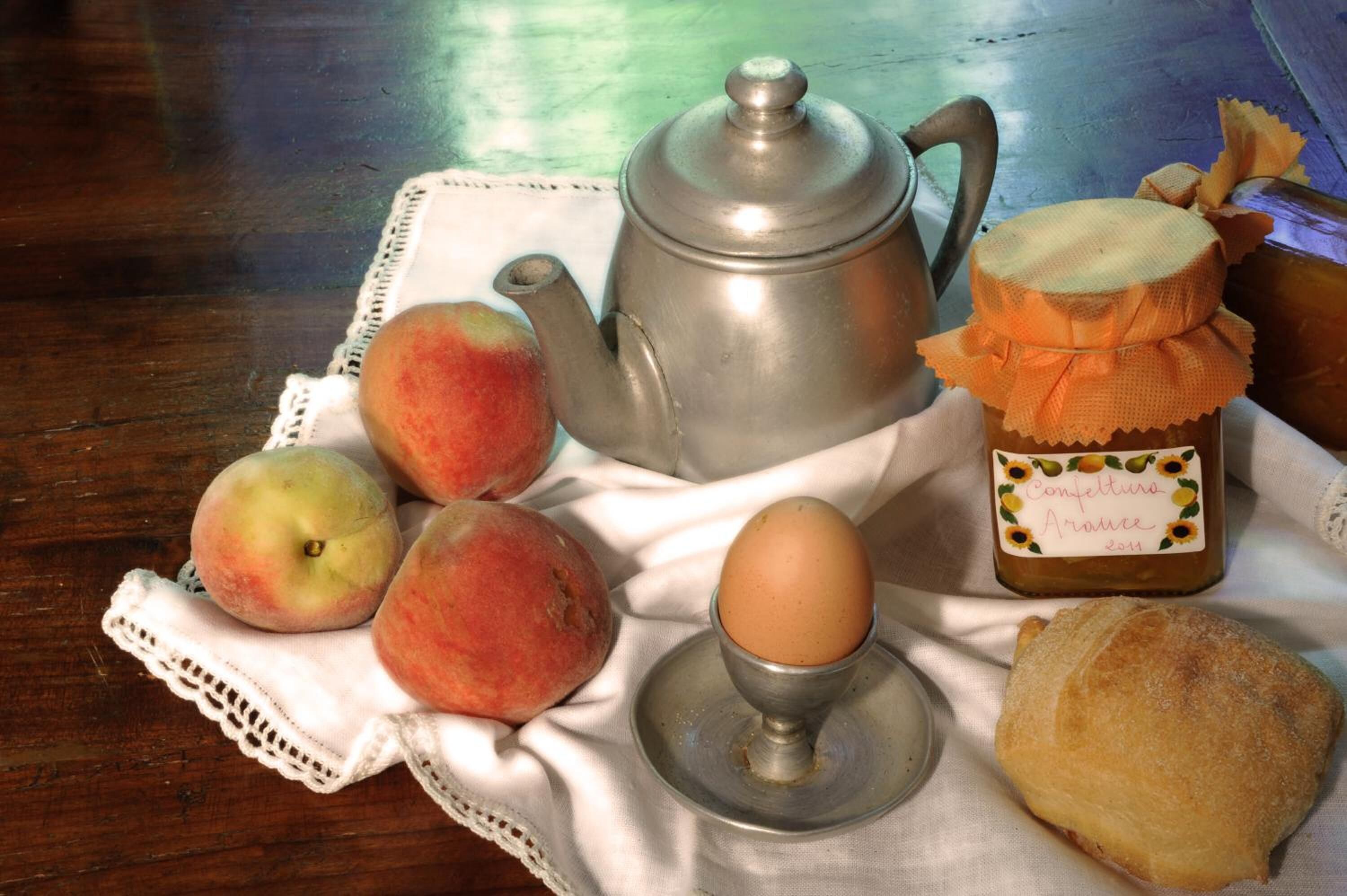 Una tavola imbandita con un servizio da tè, pesche, un uovo, un vasetto di marmellata e una pasticceria.