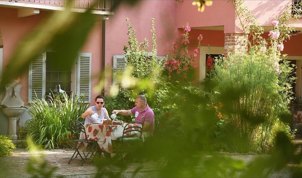 Un patio al aire libre con dos personas sentadas en una mesa, rodeado de abundante vegetación y un edificio rosa al fondo.