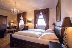 Habitación de hotel con una cama grande, un escritorio con silla, una araña y una ventana con cortinas.