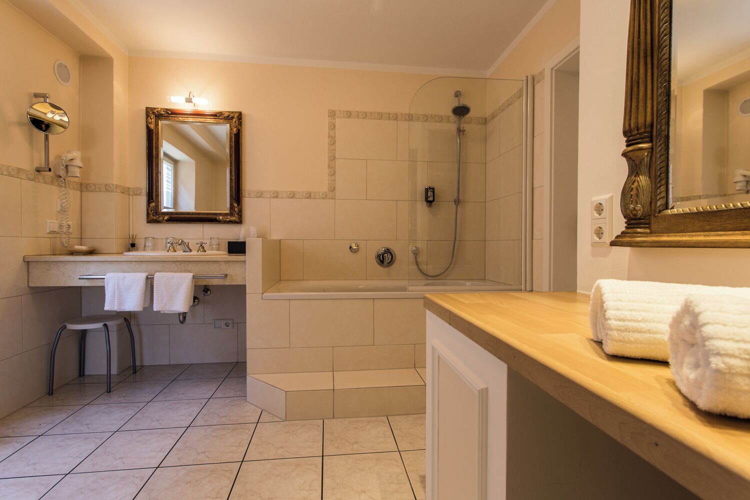 A bathroom with a bathtub, shower, vanity with a mirror, and a stool.