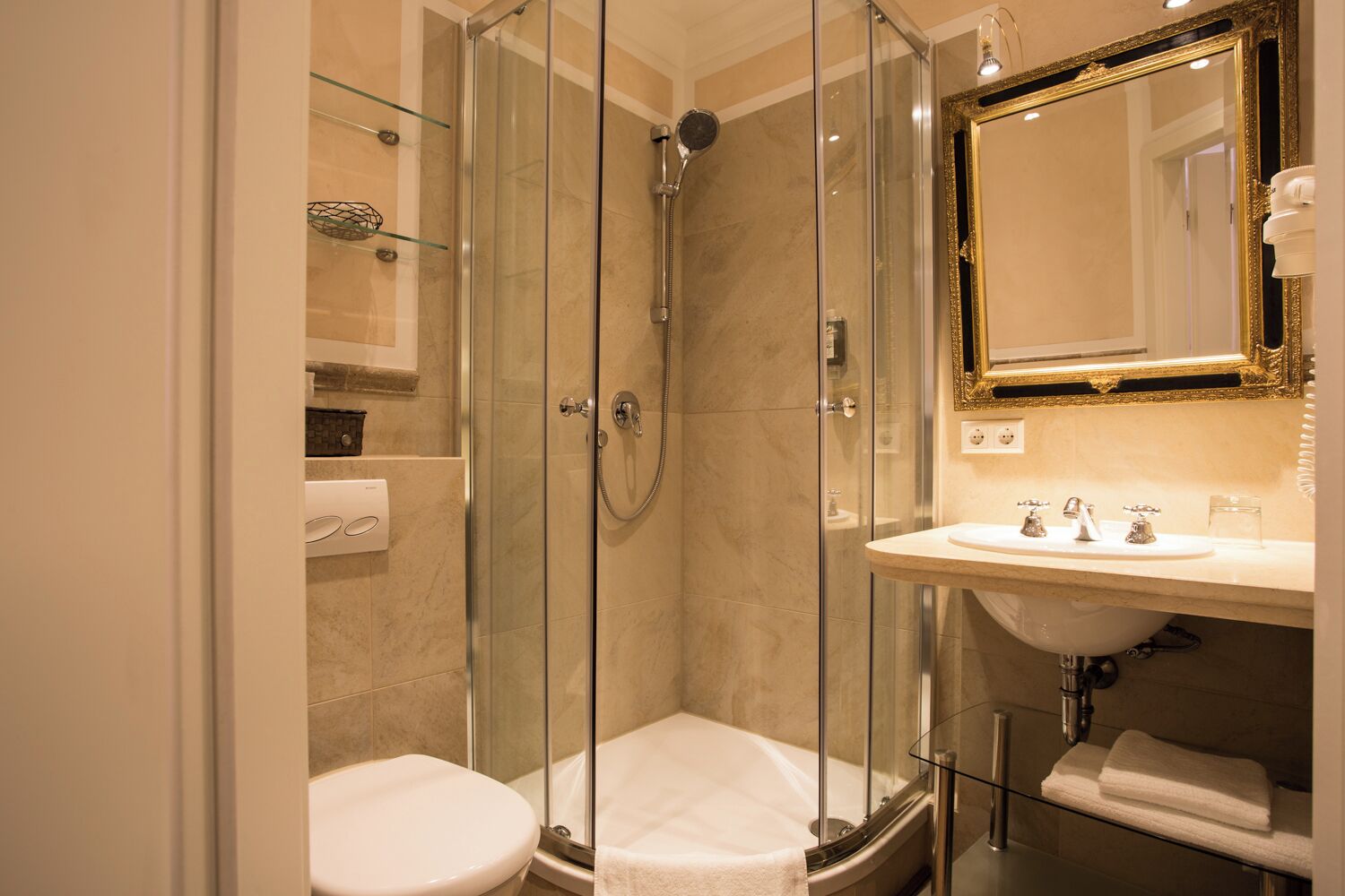 A modern bathroom with a glass-enclosed shower, a white toilet, a sink with two faucets, and a mirror with a gold frame.