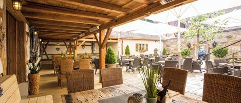 Un patio al aire libre con mesas de madera y sillas de mimbre, paredes de piedra y plantas en macetas.