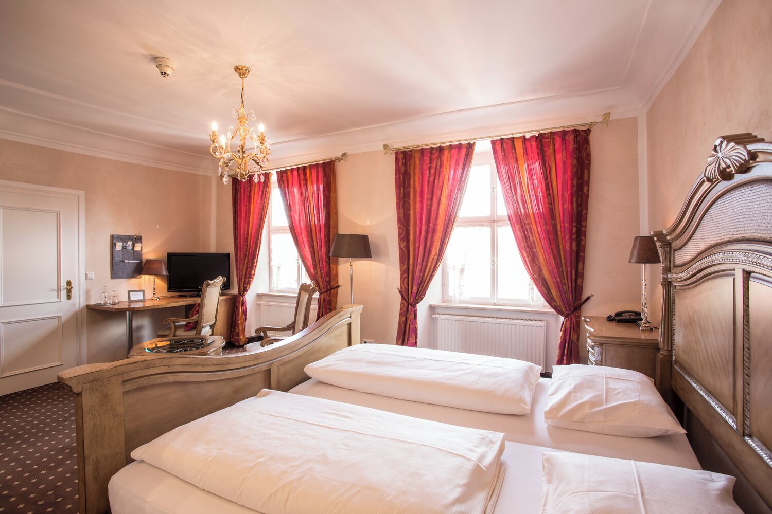 A hotel room with two beds, a desk with a computer, a chandelier, and red curtains.