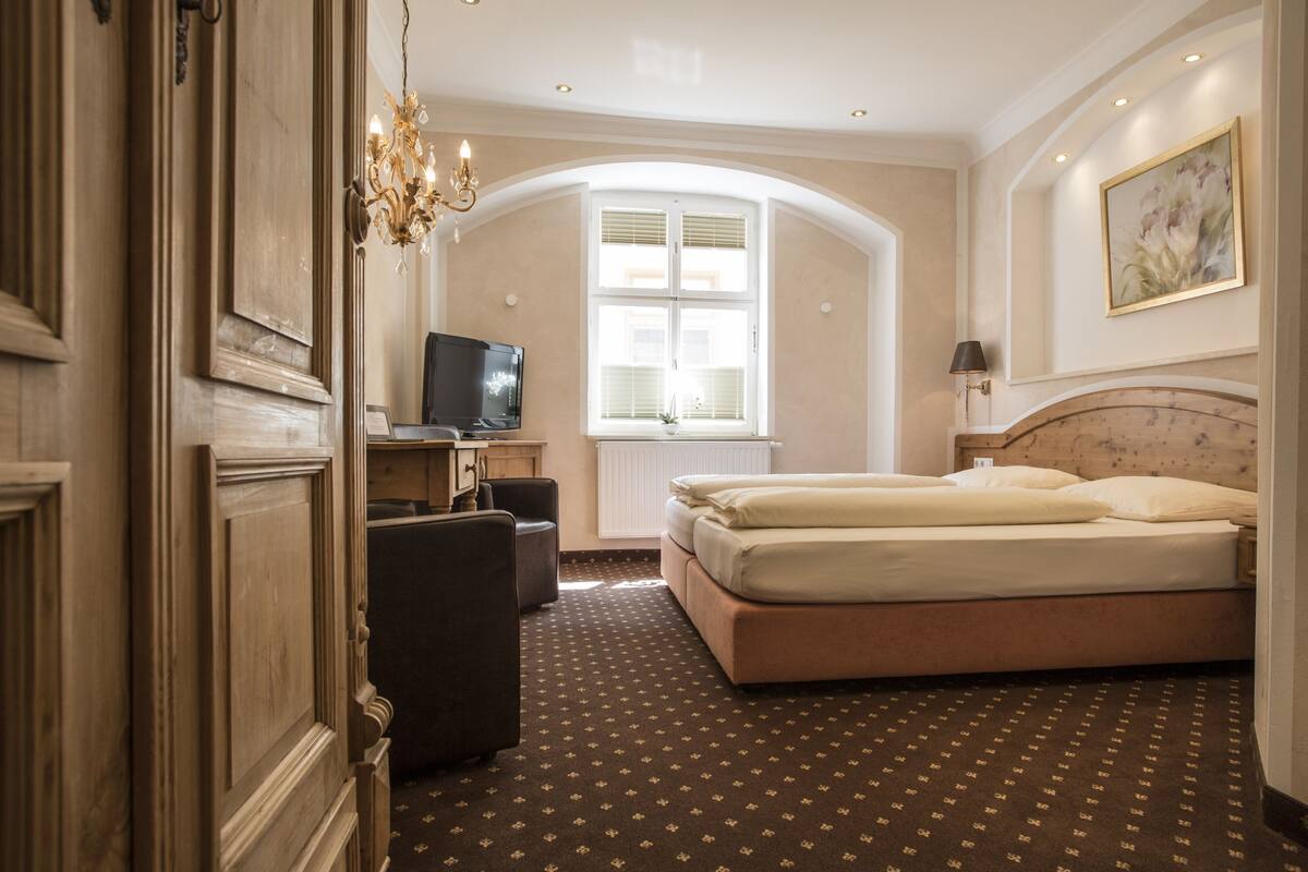Quarto de hotel com cama, televisão, escrivaninha, cadeira, lustre e janela.