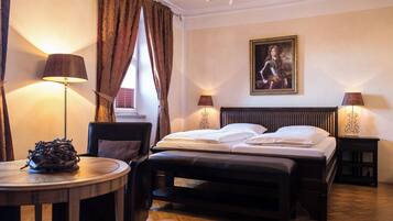 A hotel room with a bed, bedside tables, a chair, a round table, a chandelier, and a painting on the wall.