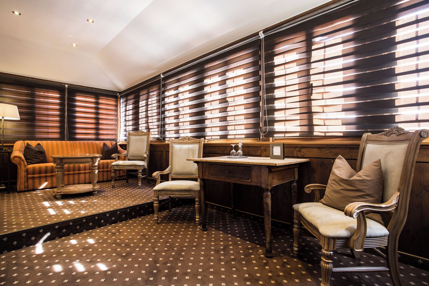 A room with a patterned floor, a sofa, two armchairs, a table, and a bookshelf with slatted shelves.