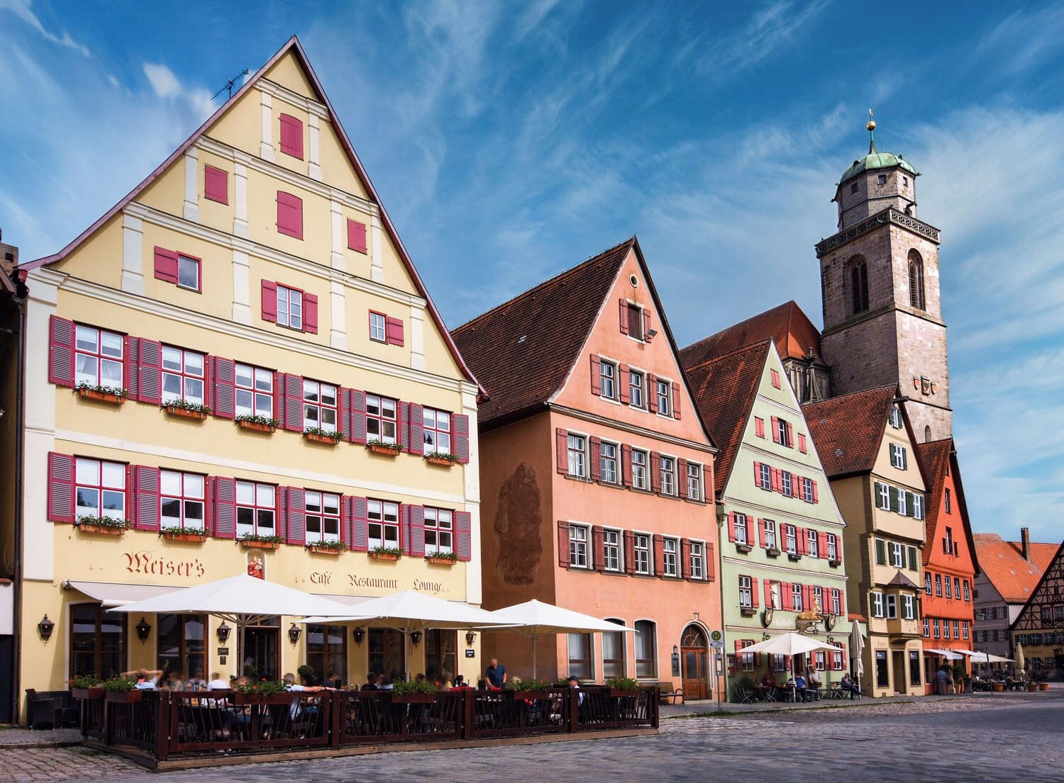 A traditional European street with colorful buildings, a prominent clock tower, and outdoor seating at a café.