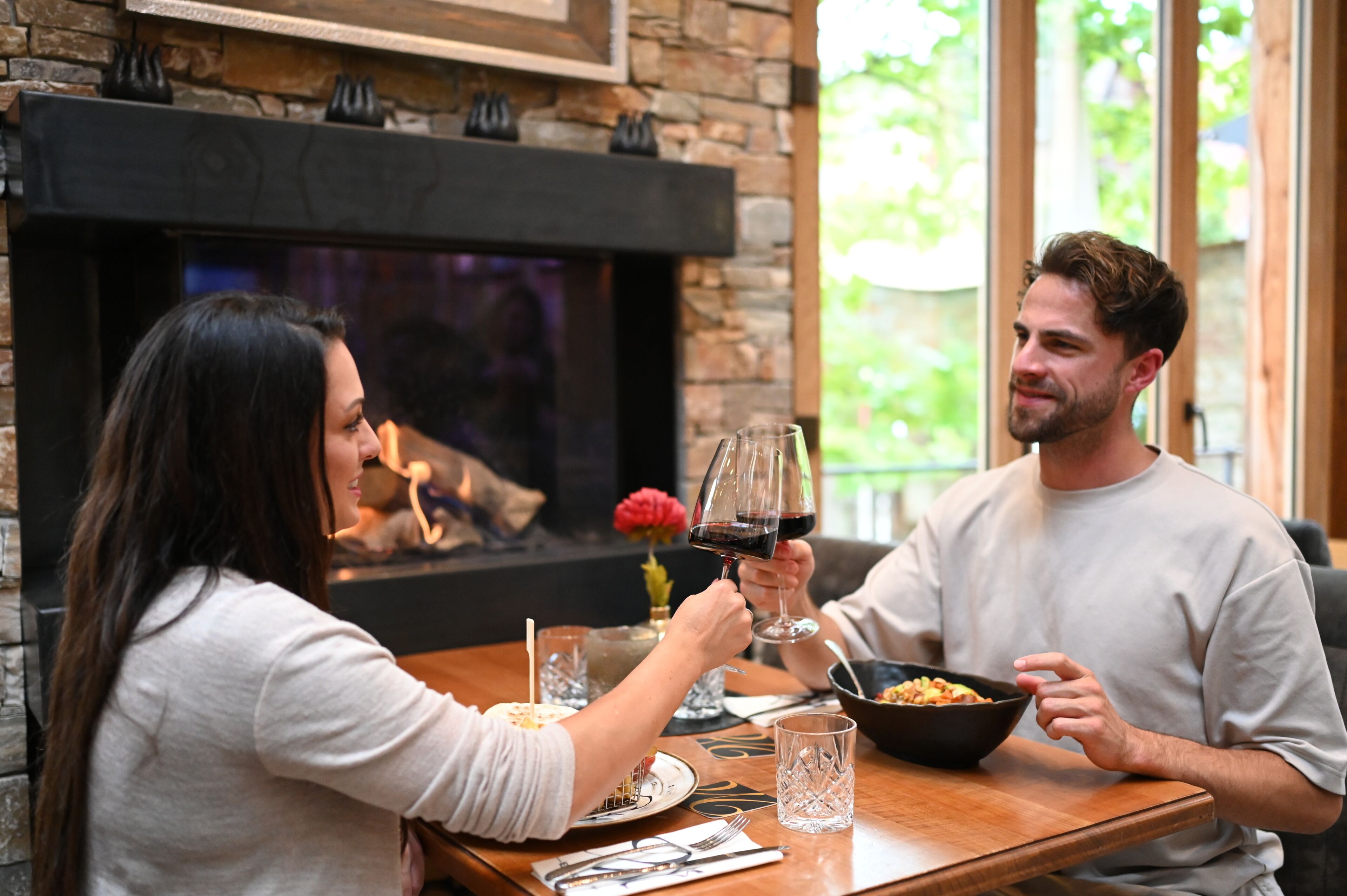Una pareja sentada en una mesa de madera con chimenea, un tazón de comida y copas de vino.
