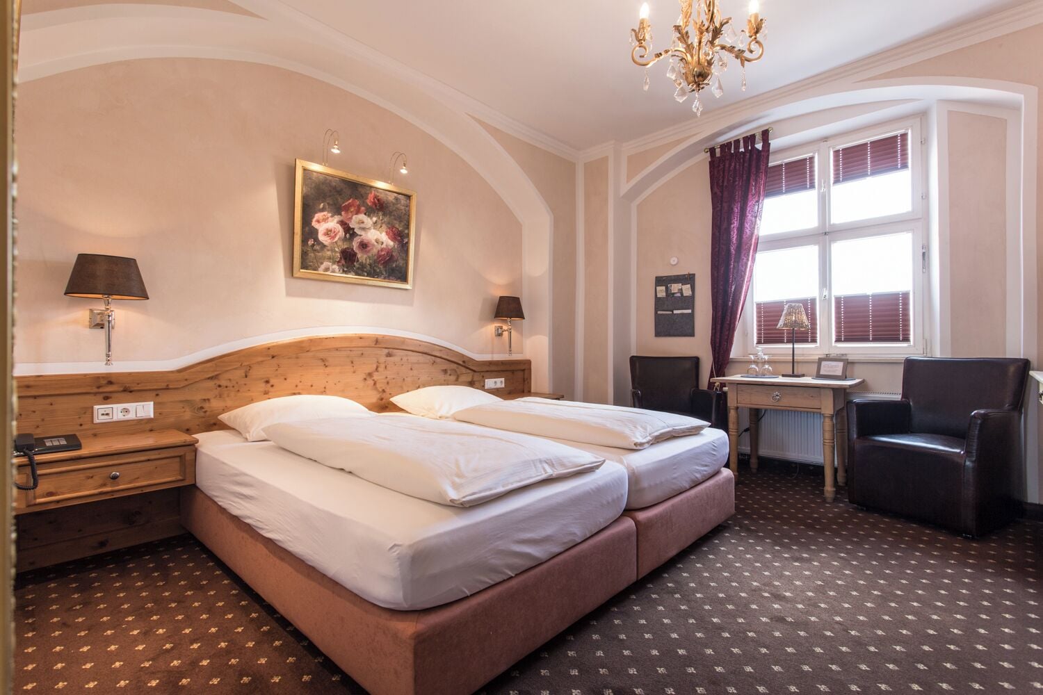 A hotel room with a double bed, a desk with a chair, a chandelier, and a window with curtains.