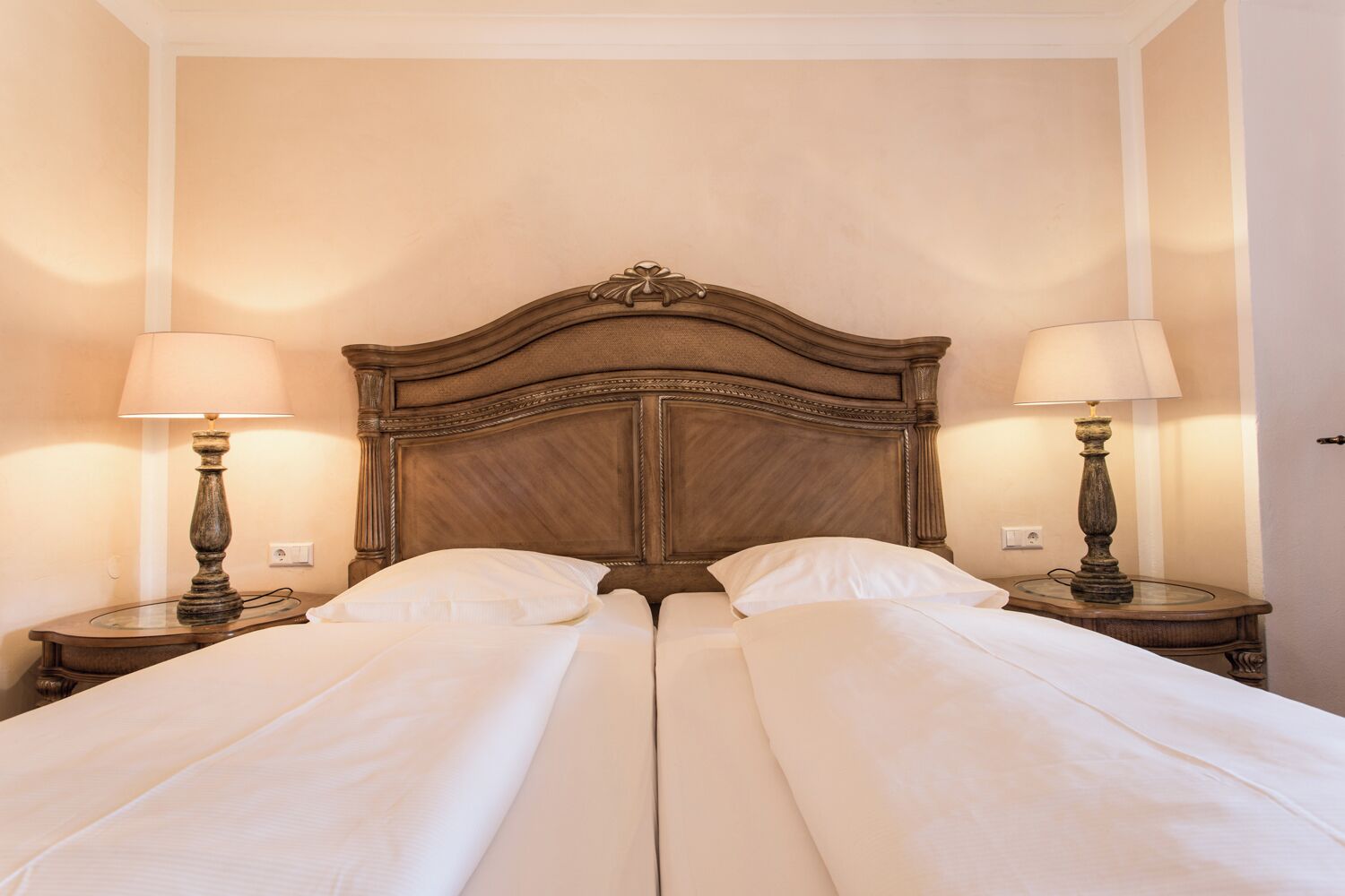 Cama doble con cabecera de madera, dos lámparas de noche y una pared lisa.